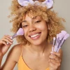 Beautiful smiling woman with curly hair applies blush on face with cosmetic brush powders cheeks looks happily at camera wears t shirt and headband isolated over grey background. Daily makeup