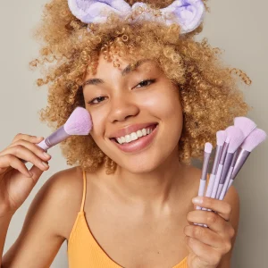Beautiful smiling woman with curly hair applies blush on face with cosmetic brush powders cheeks looks happily at camera wears t shirt and headband isolated over grey background. Daily makeup