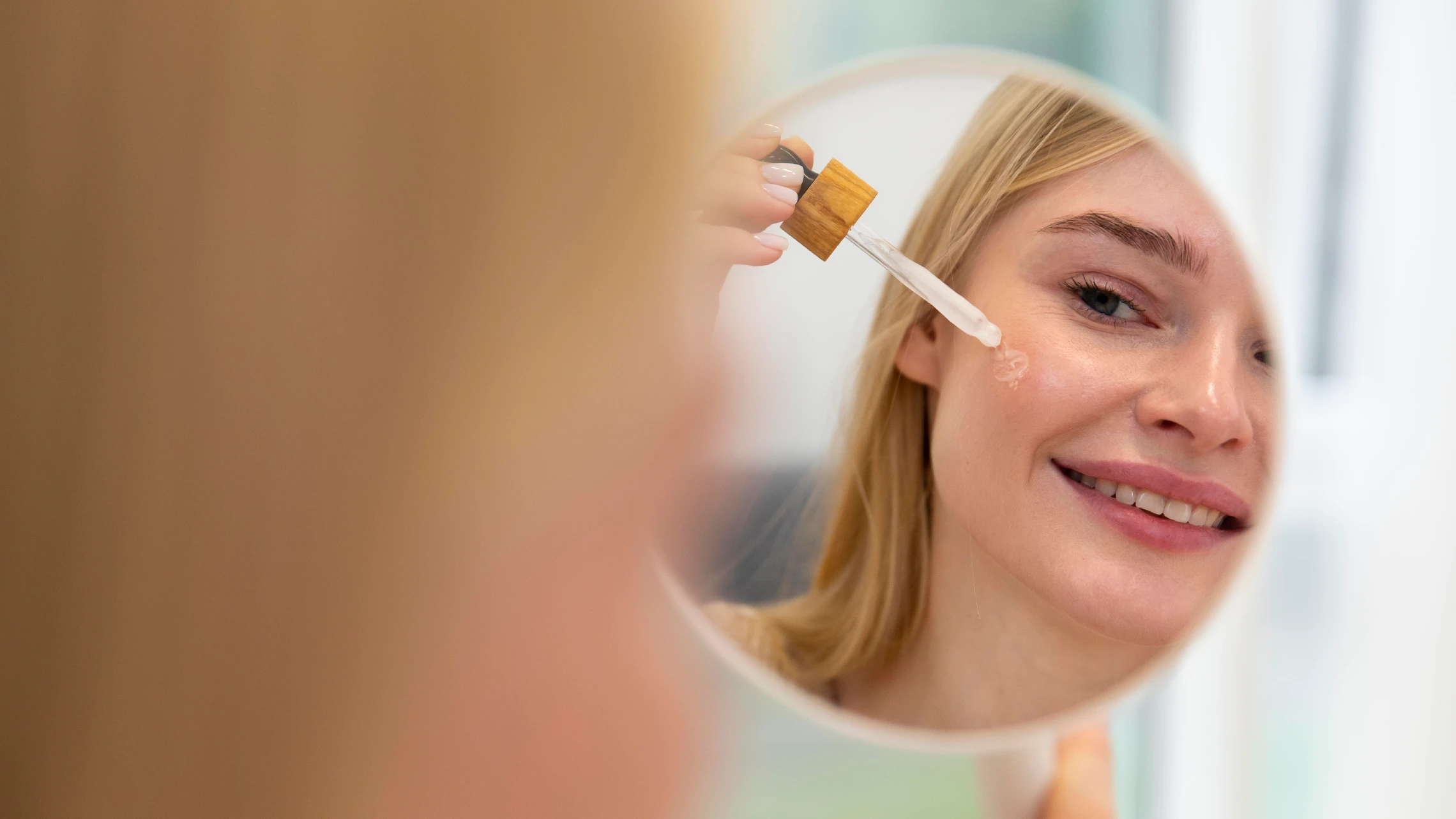 A woman smiling in a mirror reflection while applying a botanical facial oil from a dropper bottle to her cheek.