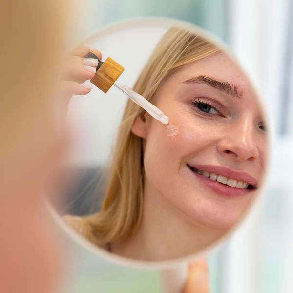 A woman smiling in a mirror reflection while applying a botanical facial oil from a dropper bottle to her cheek.