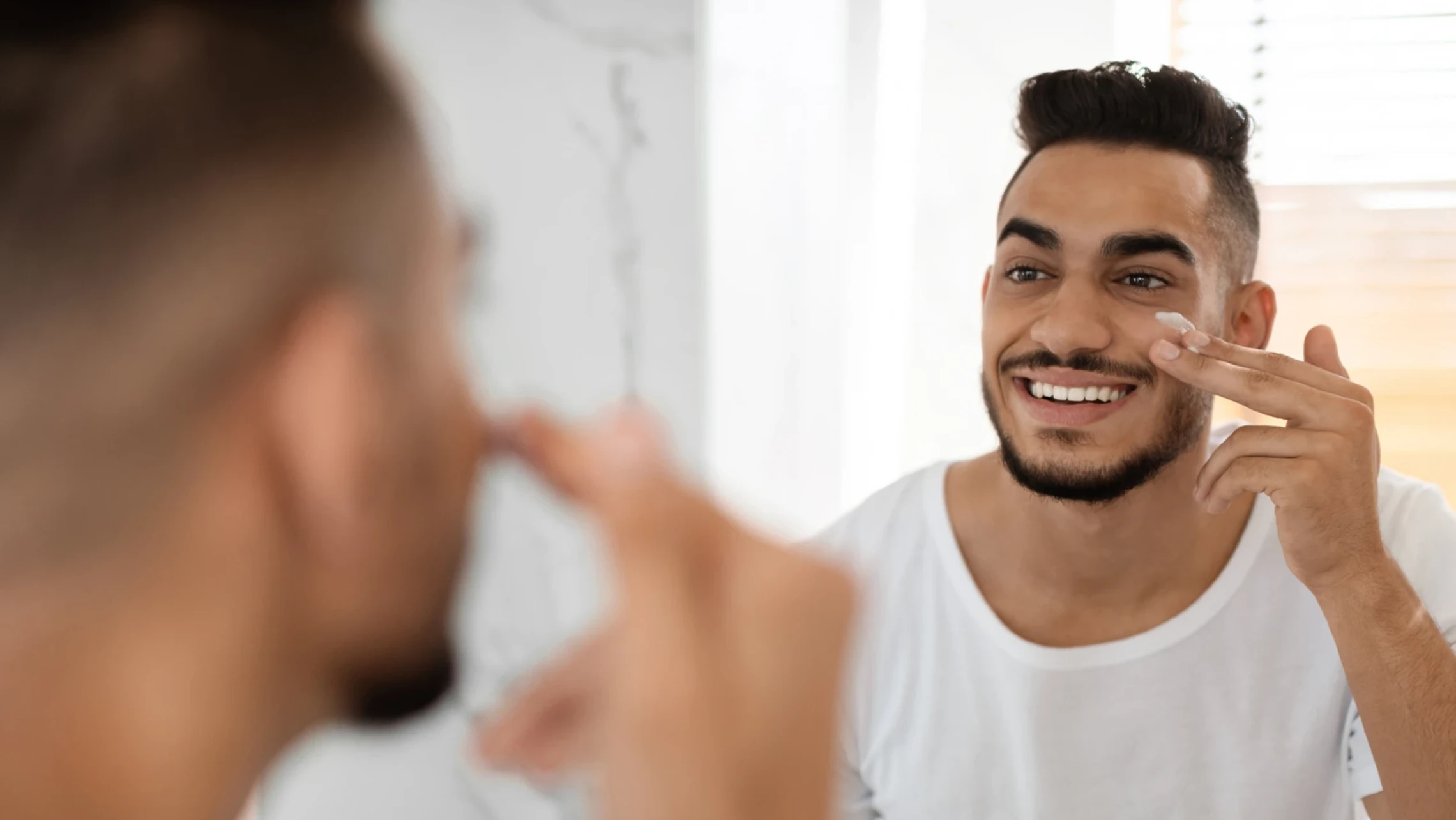 Smiling man in a bathroom mirror gently applying moisturizer cream to his cheek illustrating how to build a healthy skin barrier