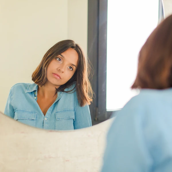 A stressed and tired woman looking at her reflection in a bathroom mirror, illustrating how chronic stress and cortisol affect skin health.