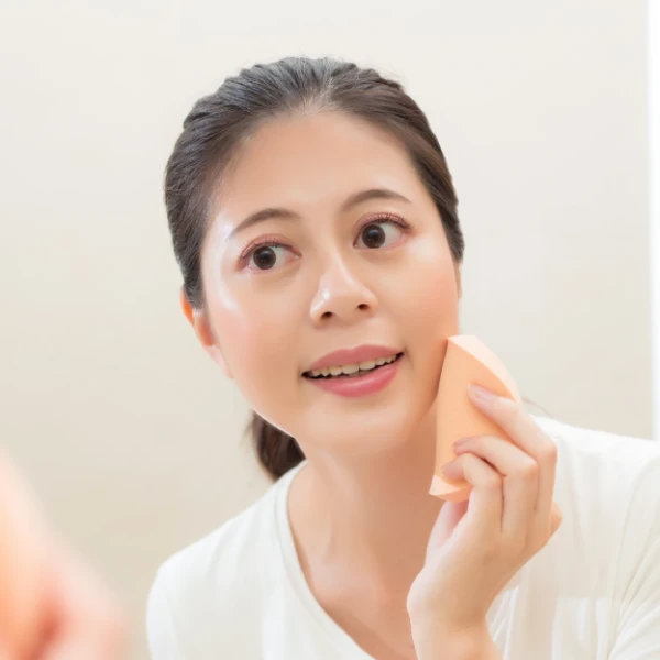 A smiling woman applying mineral makeup with a makeup sponge in the bathroom mirror, focusing on skincare tool hygiene for oily skin.